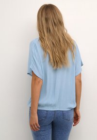 Hellblaues Kurzarmoberteil mit strukturierter Oberfläche und elastischem Saum. Kombiniert mit dunkelblauen Jeans, von hinten betrachtet.