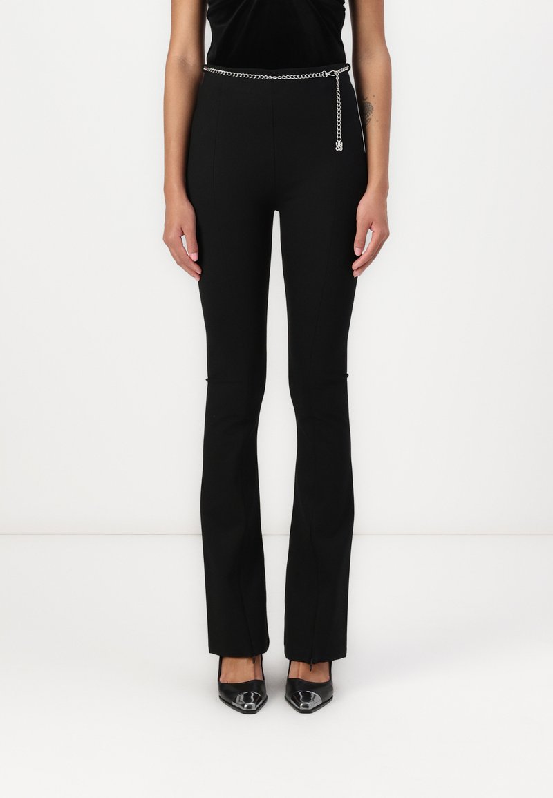 Pantalones negros de corte entallado con un diseño acampanado, que cuentan con un cinturón de cadena plateada y una textura suave. Combinados con zapatos negros de punta.