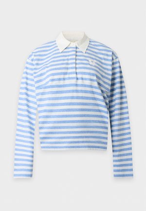 Femme debout portant une chemise à manches longues rayée bleu et blanc avec un col blanc et un jean bleu à jambes larges avec de grands pois.