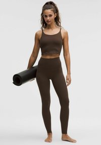 Top corto deportivo marrón y leggings de talle alto hechos de tela suave y elástica. La figura sostiene una esterilla de yoga negra enrollada bajo un brazo.