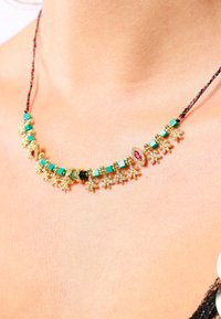 Collier en or avec des perles en turquoise et en émeraude, des accents floraux et une pierre précieuse rouge en son centre, le tout sur un cordon tressé.
