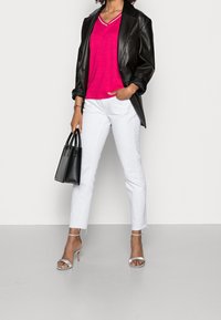 Schwarze Lederblazer, leuchtend pinkes V-Ausschnitt-Pullover, weiße Skinny-Jeans mit fransigem Saum, schwarze Handtasche und silberne Riemchen-High Heels.