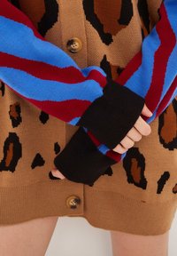 Händerna är korsade över en brun leopardmönstrad cardigan med stora knappar, och en tröja med blå och röda randiga ärmar och svarta manschetter.