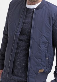 Námořnická modrá prošívaná bomber bunda se zapínáním na zip, žebrovaným límcem a vyšitým logem. Doplněna o tečkovanou tmavou košili.