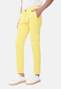 Chino in cotone giallo con vestibilità slim, tasche laterali e chiusura a bottone nero, abbinati a sneakers bianche e una camicia bianca basic.