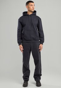 Musta huppari, jossa on kengurutasku ja säädettävä naru, yhdistetty tummanharmaisiin housuihin. Malli seisoo neutraalin taustan edessä.