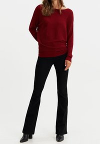 Pull rouge à côtes avec une coupe ample, encolure bateau ; associé à un pantalon noir  évasé et des bottines noires, sur fond blanc.