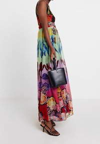 Robe maxi à motif floral aux couleurs vibrantes, avec un corsage ajusté et une jupe fluide. Associée à un petit sac à main noir et des talons à brides.