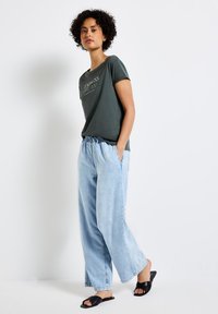 Mörkgrön kortärmad t-shirt med vit text, kombinerad med ljusblå lösa denimbyxor och svarta slidesandaler. Avslappnad outfit, ledig passform.