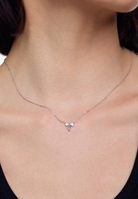 Srebrny naszyjnik z delikatnym wzorem kokardy i małymi dekoracyjnymi detalami, umocowany na cienkim łańcuszku spoczywającym na obojczyku.