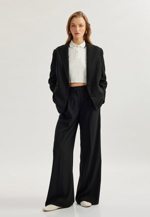Zwarte op maat gemaakte blazer over een witte cropped polo, gecombineerd met zwarte wijde broek. Opvallende details zijn de oversized pasvorm en de zachte textuur.