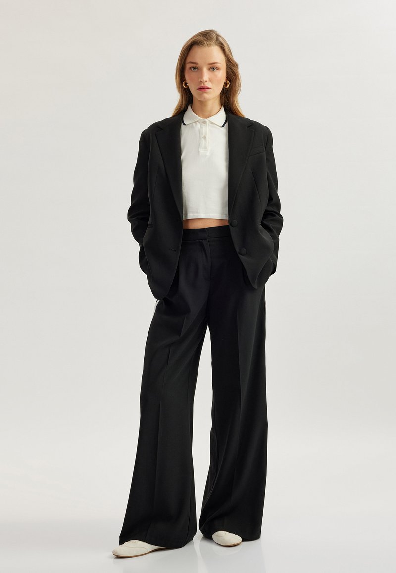 Zwarte op maat gemaakte blazer over een witte cropped polo, gecombineerd met zwarte wijde broek. Opvallende details zijn de oversized pasvorm en de zachte textuur.