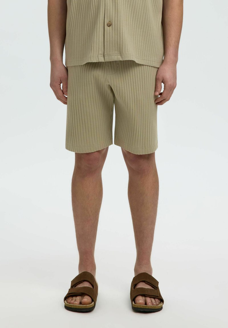 Homme portant un short côtelé beige, une chemise boutonnée assortie et des sandales marron, debout devant un fond clair uni.