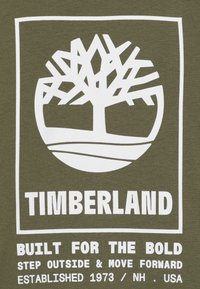 Olijfgroene katoenen t-shirt met een witte afbeelding van een boom in een cirkel en teksten: "TIMBERLAND," "GEBOUWD VOOR DE DAPPERE," en oprichtingsinformatie.