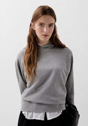 Felpa grigia con cappuccio in tessuto morbido, dalla vestibilità comoda, con tasca frontale e un discreto dettaglio del logo. Indossata sopra una camicia bianca con colletto.