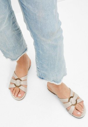 Sandales à glisser beiges avec un design à lanières croisées et un accent circulaire. Semelles texturées et coupe plate, associées à un jean bleu clair effiloché.