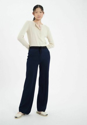Haut henley beige à manches longues avec des boutons ; pantalon navy à jambes larges avec taille élastique ; chaussures crèmes à enfiler avec couture décorative.