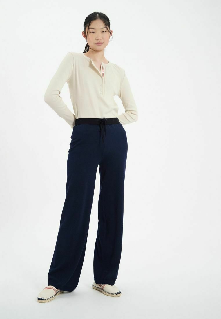 Haut henley beige à manches longues avec des boutons ; pantalon navy à jambes larges avec taille élastique ; chaussures crèmes à enfiler avec couture décorative.