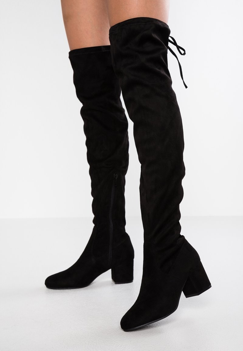 Bottes hautes en daim noir au-dessus du genou avec un talon bloc épais, dotées d'une fermeture éclair latérale et d'un détail de lacets en haut. Texture lisse.