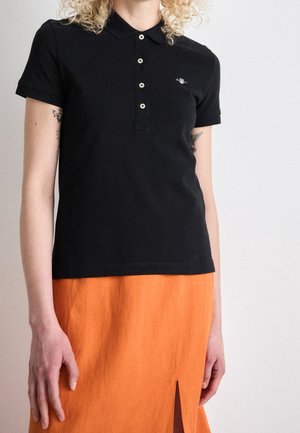 Personne portant un polo noir à manches courtes avec des boutons et une jupe orange fendue à l'avant. Tatouages visibles sur les bras supérieurs.