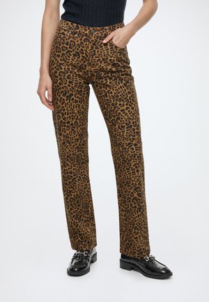 Pantalon marron droit avec des taches léopard noires, porté avec des mocassins noirs et un haut noir sans manches, une main dans la poche.