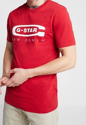 Mężczyzna w czerwonym t-shircie G-Star Raw Denim i beżowych spodniach, z widocznym tatuażem na szyi, z rękami złożonymi przed sobą.