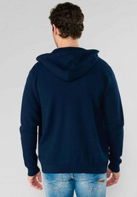 Navyblauwe pullover hoodie met een zachte textuur, vaste capuchon en geribbelde manchetten, gedragen over lichtblauwe denim jeans.