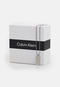 Sølv kjede med rektangulært vedheng, med graverte linjer, hviler på en hvit boks med en svart stripe og "Calvin Klein"-logo.