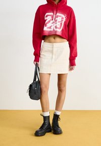Felpa rossa corta con grafica bianca, mini gonna a righe crema, stivali neri con lacci e una piccola borsa nera. Superficie piatta sullo sfondo.