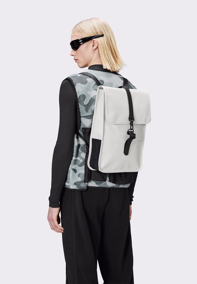 Vit läderryggsäck med lock, svart rem och sidoficka, kombinerad med en nät-camouflageväst och svart långärmad skjorta.