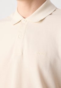 Polo beige avec deux boutons et un petit logo BOSS brodé sur la poitrine gauche, porté par une personne sur un fond uni.