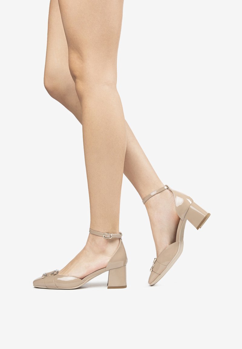 Beige patent leather block heel shoes with ankle strap, featuring a square toe and decorative buckle accents. Smooth texture, minimalistic design.