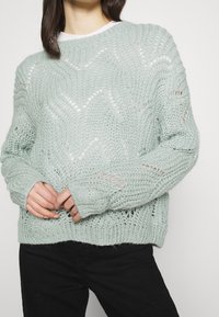 Pull en maille vert menthe avec un tissage lâche et ouvert, présentant un motif en zigzag et des poignets côtelés, porté par-dessus un haut blanc.