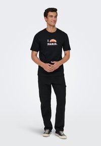 Μαύρο βαμβακερό μπλουζάκι με σχέδιο κρουασάν και το κείμενο "I PARIS." Συνδυασμένο με μαύρο παντελόνι και καθημερινά παπούτσια.