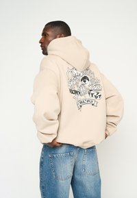 Sweatshirt beige avec un grand design graphique noir au dos, comportant divers éléments de texte et un détail d'œil. Texture en mélange de coton.