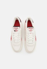 White sneakers with red accents, lace-up design, and perforations on the toe box. Features a black and red logo on the tongue.
