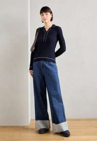 Top de manga larga de nailon de color marino con cuello y tapeta, combinado con jeans de pierna ancha de mezclilla que presentan un dobladillo sin terminar y acentos en gris claro.