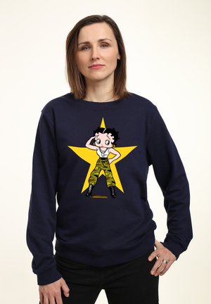 Mørkeblå sweatshirt med en tegneseriefigur iført militærgrøn camo-bukser, sorte støvler og en hvid tank top, sat mod en gul stjerne.