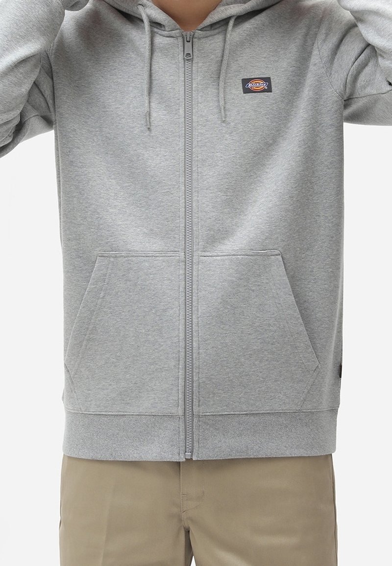 Personne portant un sweat à capuche gris clair avec fermeture éclair, poches avant et petit écusson logo, assorti à un pantalon beige.