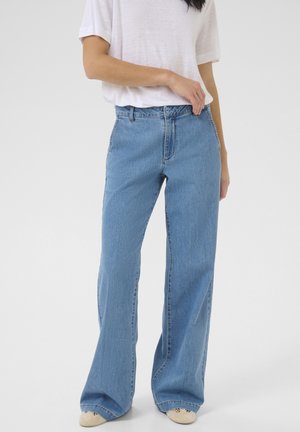 Personne portant un jean bleu large, un t-shirt blanc à manches courtes et des chaussures beiges, debout devant un fond clair uni.