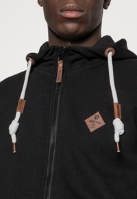 Sort zip-up hættetrøje lavet af blødt stof. Har et lædermærke, hvide snore og metal detaljer på lynlåsen.