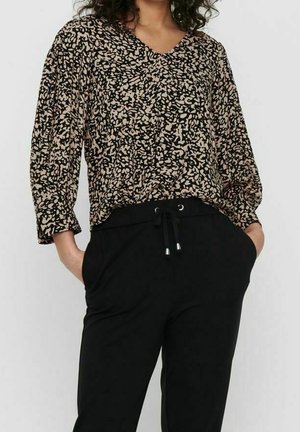 Frau trägt eine langärmelige, gemusterte beige und schwarze Bluse mit schwarzen Kordelhosen, Hände lässig in den Taschen.