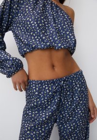 Zkrácený top s modrým květinovým vzorem a jedním rukávem; odpovídající kalhoty na šňůrku. Materiál vypadá lehký a má měkkou strukturu.