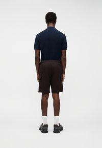 Άντρας που φοράει μπλε κοντομάνικο πόλο, μαύρες σορτς cargo, άσπρες κάλτσες και μαύρα αθλητικά παπούτσια, όρθιος με την πλάτη στραμμένη προς την κάμερα.