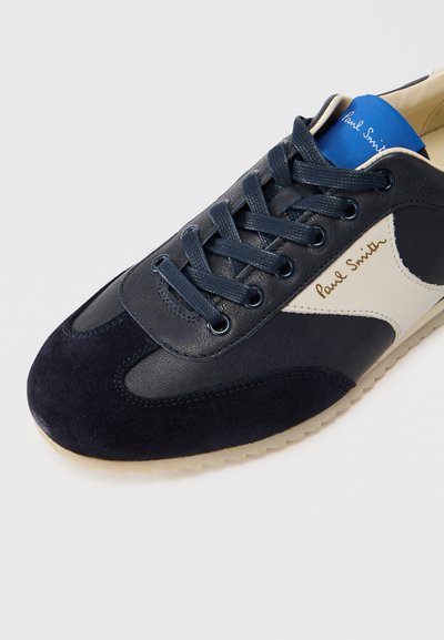 Σκούρο μπλε σουέντ και δερμάτινο αθλητικό παπούτσι με εκρού πλαϊνή επιφάνεια, μπλε εσωτερικό και επίπεδη λαστιχένια σόλα. Διαθέτει στρογγυλά κορδόνια και χρυσή λεπτομέρεια με λογότυπο.