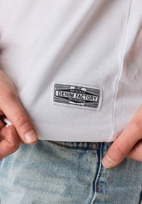 Valkoinen puuvillapaita, jossa on suorakulmainen musta etiketti, jossa lukee "DENIM FACTORY" sivussa. Yhdistä vaaleansiniset kulutetut farkut.