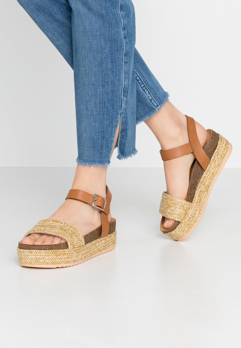 Bruna remsandaler med en flätad juteplattform och korkbas. Designen har överdimensionerade korsremmar och en justerbar ankelspänne.