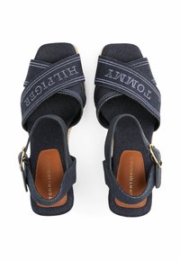 Navy fabric sandals with crossed straps featuring 'Tommy Hilfiger' text. Buckle closure, textured surface, and a brown leather insole.