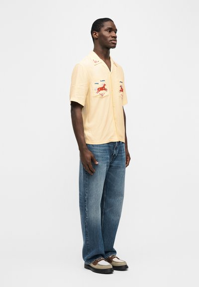 Άνδρας που στέκεται, φοράει ανοιχτό κίτρινο πουκάμισο με κοντά μανίκια και κόκκινη κεντημένη εικόνα αλόγου, μπλε τζιν και καφέ-μπεζ δερμάτινα παπούτσια με κορδόνια.