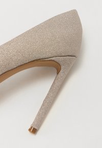Scarpa con tacco alto beige glitterato, dalla punta affusolata, con un sottile tacco a stiletto e un interno in pelle liscia. Presenta una superficie testurizzata e luccicante.